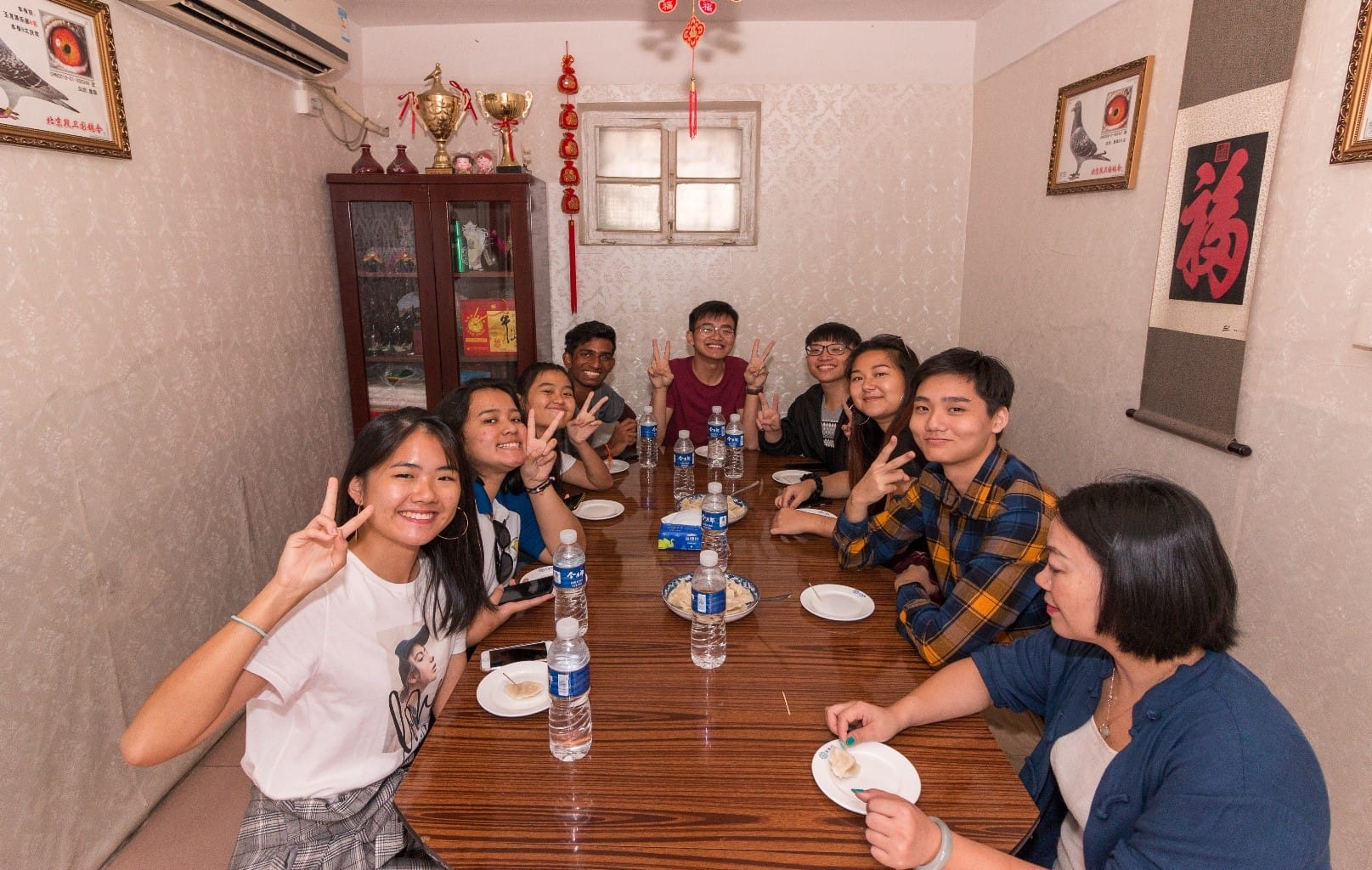 A group of young people sit around a wooden dining table, smiling and making peace signs in a decorated room.